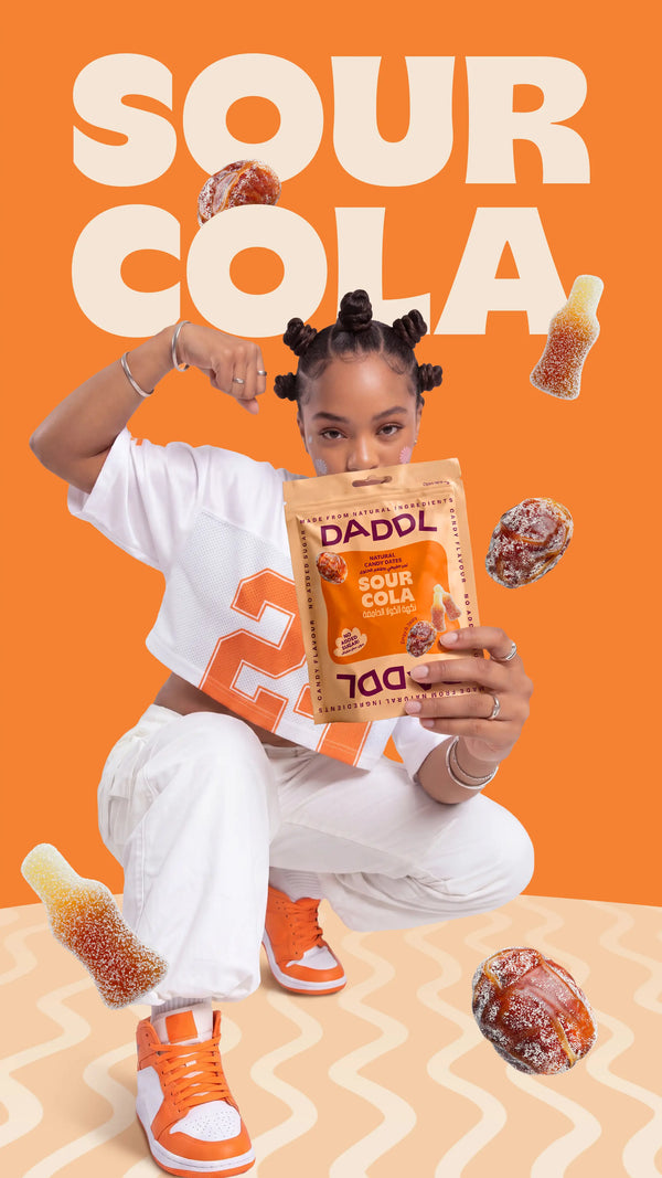 Young woman in white and orange streetwear crouching while holding a DADDL Sour Cola candy dates pouch against a bright orange background with large “SOUR COLA” text, floating coated dates, and cola bottle gummy candies.