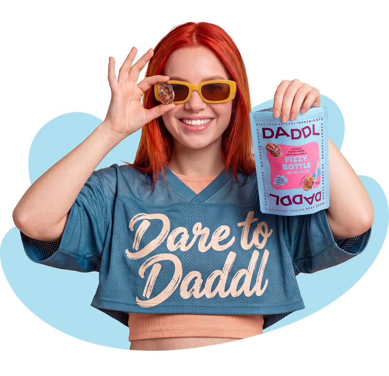 Smiling woman with bright red hair wearing yellow sunglasses and a blue “Dare to Daddl” shirt while holding a DADDL Fizzy Bottle candy dates pouch and a coated date in front of her eye against a transparent background with abstract blue shapes.