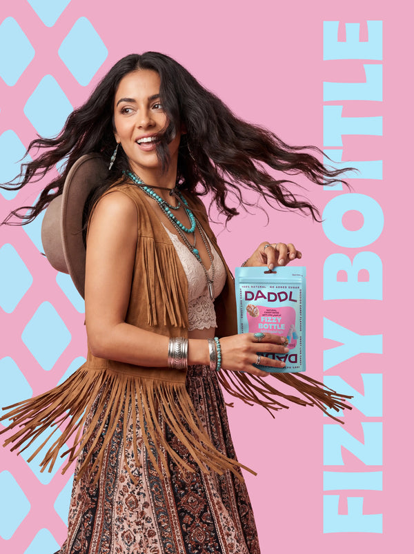 Woman dancing while holding DADDL Fizzy Bottle natural date candy pack on a pink background with bold graphic patterns.