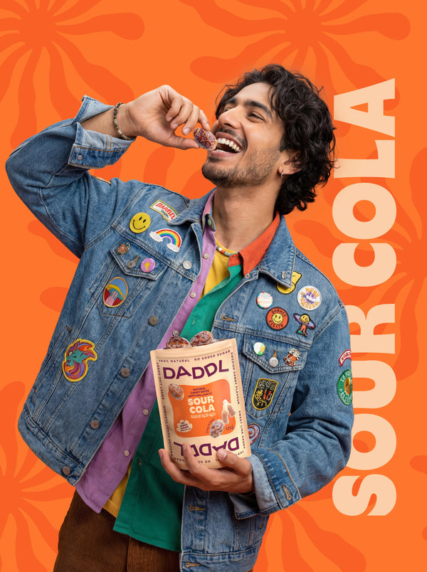 Man eating a date candy while holding DADDL Sour Cola pack on an orange graphic background.