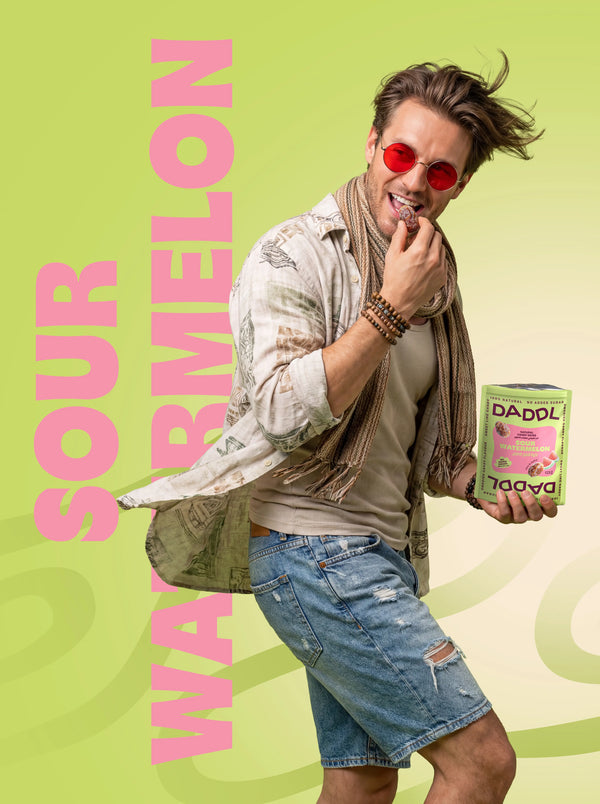 Man smiling and eating a date candy while holding DADDL Sour Watermelon pack on a green background.